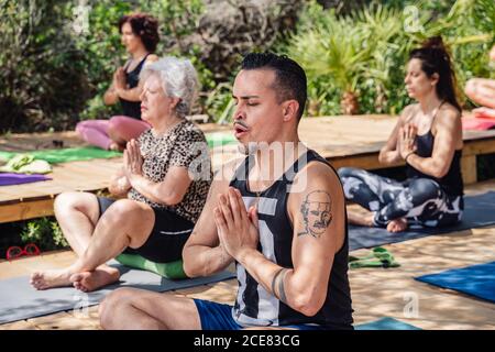 Junger Mann und ältere Frau in einer Gruppe von Menschen meditieren in Lotus-Pose mit geschlossenen Augen während Yoga-Praxis in tropischen Resort an sonnigen Tag Stockfoto