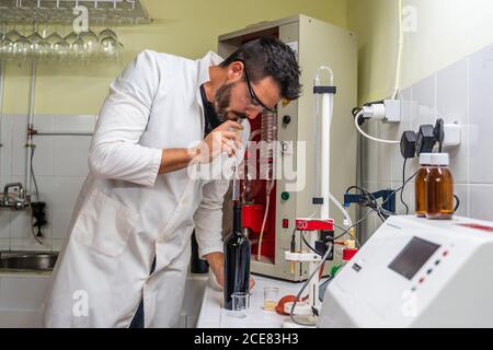 Erwachsene männliche Wissenschaftler in Uniform und Brillen, die den Zuckergehalt messen Des Weins bei Verwendung eines transparenten Weinzählers im Laborbetrieb In der Nähe von elektronischen Geräten Stockfoto