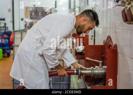 Seitenansicht ernsthafte Winzer in weißem Gewand Befestigung flexible Rohr Zum Fermentationstank während der Arbeit in zeitgemäßer professioneller Fabrik Stockfoto