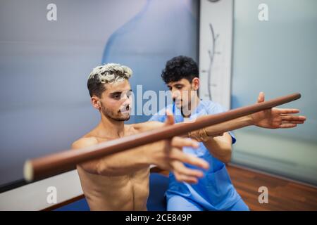 Von oben des jungen Osteopathen in der Uniform helfenden muskulären Menschen Mit gefärbten Haaren halten Holzstab mit erreichten Armen während Wegschauen Stockfoto