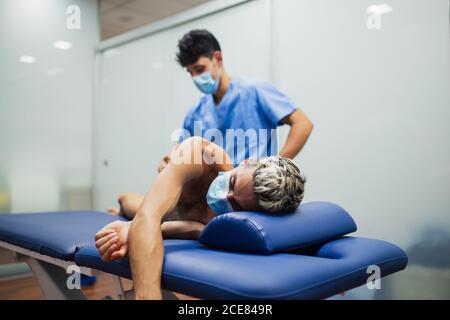 Osteopath mit Maske in blau Uniform Untersuchung Rückseite schlank Männlicher Patient mit Maske und gefärbtem Haar liegend auf Untersuchung Tisch in der Klinik Stockfoto