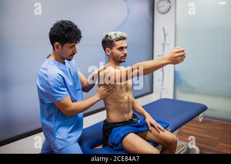 Seitenansicht des Orthopäden bei gleichmäßiger Überprüfung des Schultergelenks Der fitting Mann sitzt auf der Untersuchungstisch mit erreichten Arm Im Krankenhaus und wegschauen Stockfoto