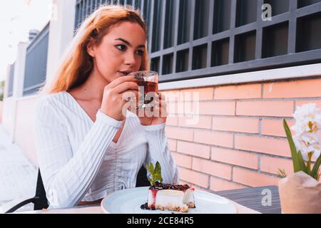 Junge verträumte Dame, die heißen Tee trinkt und dabei wegschaut Sitzen am Holztisch mit leckeren Heidelbeer Käsekuchen dekoriert mit Frische Minze in der Nähe der Ziegelmauer in der Cafeteria im Hinterlicht Stockfoto