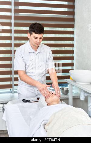 Männliche Kosmetikerin spendet Feuchtigkeit und reinigt das Gesicht der Frau mit Sahne und Seife. Spa-Behandlungen Stockfoto