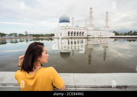 Rückansicht der ethnischen weiblichen Touristen lehnt an Grenze in der Nähe see vor der berühmten Bandaraya Moschee und Blick weg Während Sommerferien in Kota Kinabalu Stockfoto