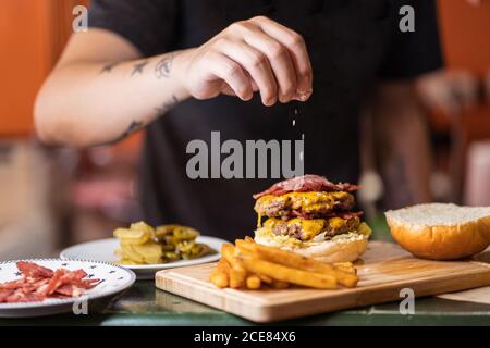 Nicht erkennbarer männlicher Koch, der beim Aufstehen Salz zum Burger hinzufügt Tisch im Café Stockfoto