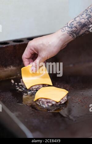 Unkenntlich männlicher Koch mit Tattoo Zugabe von Scheiben Käse auf Cutlets beim Zubereiten von Burgern im Café Stockfoto