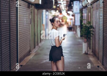 Ganzkörper junge asiatische Frau in lässiger Kleidung mit Mobile Telefon in der Hand zu Fuß auf engen Fußgänger unterirdischen Weg mit Geschlossene Geschäfte in der Stadt Stockfoto