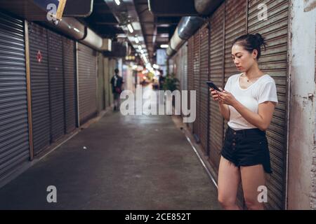 Ganzkörper junge asiatische Frau in lässiger Kleidung mit Mobile Telefon in der Hand zu Fuß auf engen Fußgänger unterirdischen Weg mit Geschlossene Geschäfte in der Stadt Stockfoto