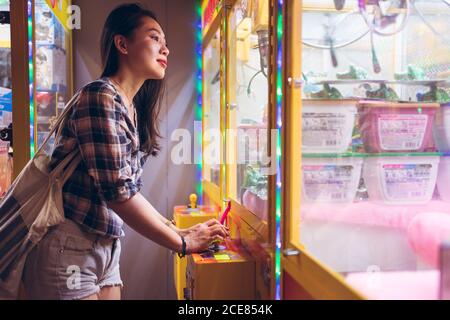 Seitenansicht der positiven jungen asiatischen Frau, die om Claw spielt Kranmaschine mit bunten Preisen Stockfoto