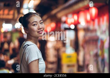 Seitenansicht von glücklich junge asiatische Frau in Freizeitkleidung Blick auf die Kamera und lächeln, während sich gegen verschwommenes Licht City Street in der Nacht Stockfoto