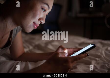 Seitenansicht der fröhlichen jungen asiatischen Frau mit Smartphone, während Nachts zu Hause im Bett liegen Stockfoto