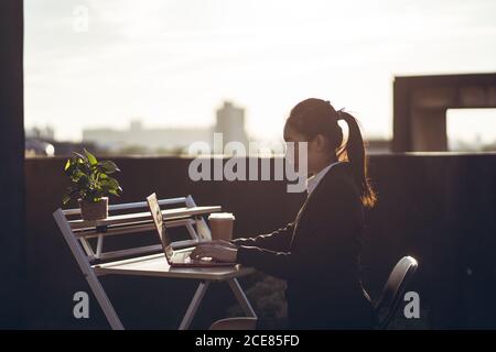 Seitenansicht der jungen asiatischen Geschäftsfrau in formellem Outfit Mit einem Laptop arbeiten, während Sie auf der Dachterrasse in der Stadt arbeiten Stockfoto