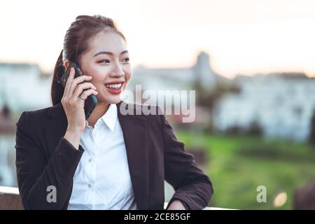 Seitenansicht der jungen asiatischen Geschäftsfrau in formellem Outfit Telefonieren auf dem Smartphone, während Sie auf der Dachterrasse arbeiten Stadt Stockfoto