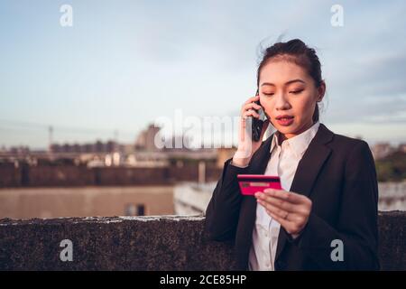 Seriöse junge asiatische Geschäftsfrau in formeller Kleidung mit Kreditkarte und Telefongespräch mit dem Bankkundendienst, während sie auf dem Dach des Stadtgebäudes steht Stockfoto