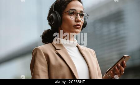 Selbstbewusste weibliche Führungskraft, die Musik auf dem Handy hört. Geschäftsfrau trägt Kopfhörer und hält ein Telefon, während sie im Freien auf Stadt stre stehen Stockfoto