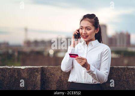 Seriöse junge asiatische Geschäftsfrau in formeller Kleidung mit Kreditkarte und Telefongespräch mit dem Bankkundendienst, während sie auf dem Dach des Stadtgebäudes steht Stockfoto