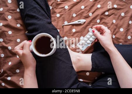 Von oben ungesund beschnitten unkenntlich weiblich sitzend auf Bett mit tasse heiße Getränke und Papiertaschentücher während der Behandlung kalt Mit Medizin Stockfoto