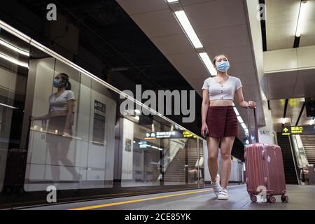 In voller Länge ruhig junge asiatische Frau in legere Kleidung und Gesichtsmaske mit Koffer bereit, moderne Schnellzug zu besteigen Kabine im modernen Bahnhof Stockfoto