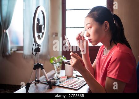 Junge asiatische Frau in Freizeitkleidung hält Spiegel und die Anwendung der Grundlage auf Gesicht, während Video-Tutorial für Beauty-Blog zu Hause Arbeitsplatz mit Ringlicht und Kamera Stockfoto