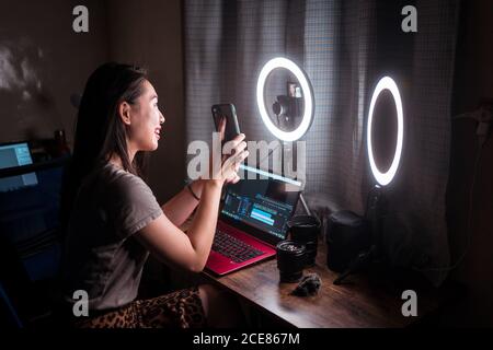 Seitenansicht einer positiven jungen asiatischen Influencerin, die modern zeigt Handy beim Sitzen am Tisch mit Gadgets und Kamera Mit Ringlicht und Video für soziale Medien Stockfoto