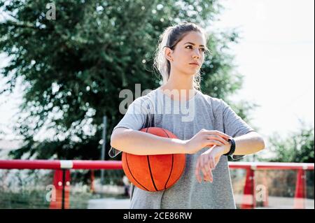Niedriger Winkel der seriösen jungen Frau in Sportbekleidung hält Basketball Ball und Kontrolle der Zeit auf der Armbanduhr während der Wartezeit auf einen Freund Zum gemeinsamen Training auf dem Platz Stockfoto