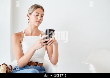 Weibliche Klientin sitzt auf einem Stuhl und browsing auf dem Smartphone während Warten auf Termin in der Lobby der modernen Klinik Stockfoto