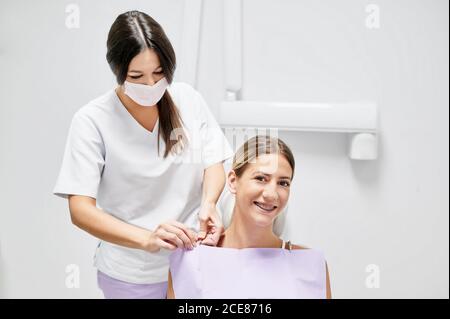 Fröhliche Patientin lächelt und schaut während des Sitzens auf die Kamera Auf dem Stuhl neben Zahnarzt während des Besuchs in der modernen Zahnklinik Stockfoto