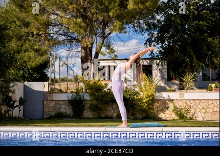 Seitenansicht der schlanken Frauen mit Sport-BH und Leggings Stehen auf Matte in der Nähe von Pool und Yoga in Urdhva üben Hastasana Stockfoto