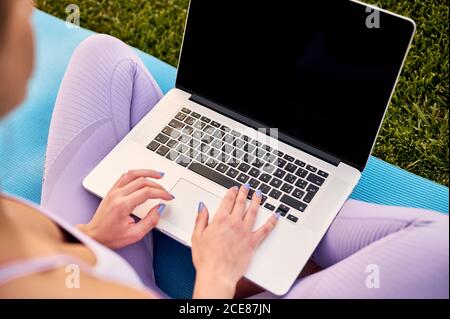Von oben der Ernte unkenntlich fit Frau in Sportkleidung sitzt mit gekreuzten Beinen auf Yoga-Matte, während Surfen im Internet auf Netbook mit schwarzem Bildschirm bei Tageslicht Stockfoto