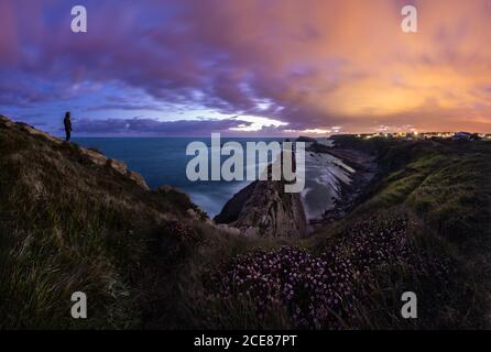 Seitenansicht Silhouette von anonymen männlichen Wanderer auf Hang stehen Von Hügel an felsiger Küste und bewundern spektakuläre Landschaft mit Farbenfroher, wolkiger Himmel bei Sonnenuntergang Stockfoto