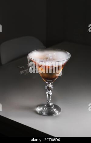 Elegantes Cocktailglas mit braunem alkoholischen Getränk und rauchend trocken Eis auf grauem Tisch Stockfoto