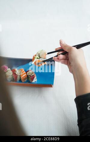 Von oben eine gesichtslose Frau, die Sushi mit Essstäbchen über dem weißen Holztisch hält Stockfoto