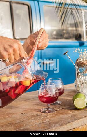Gesichtslose Person Gießen kühlen Sangria in Kristallgläsern auf Holz Tisch gegen blauen Van am tropischen Strand Stockfoto