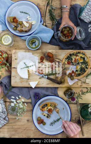 Draufsicht der Ernte unkenntlich Partner essen lecker Quiche mit Sonnengetrocknete Tomaten und Burrata Käse in der Nähe von Glas grün Oliven beim Mittagessen am Holztisch im Café Stockfoto