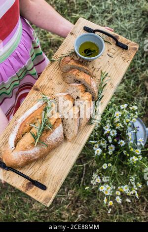 Draufsicht auf Ernte anonyme Frau mit Holz Schneidebrett Mit schmackhaftem Brot geschnitten auf Lafs mit zarter Textur verziert Mit frischen Rosmarinzweigen in der Nähe Schüssel mit Olivenöl Stockfoto