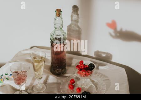 Von oben Flasche hausgemachten Rosenwein in Kristall gegossen Glas in der Nähe Champagner Trompete und serviert mit Teller frisch Himbeeren und Brombeeren gegen weiße Wand mit Handschatten Stockfoto