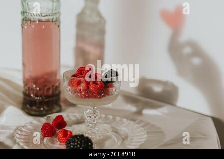 Von oben Flasche hausgemachten Rosenwein in Kristall gegossen Glas in der Nähe Champagner Trompete und serviert mit Teller frisch Himbeeren und Brombeeren gegen weiße Wand mit Handschatten Stockfoto