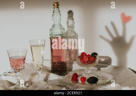 Von oben Flasche hausgemachten Rosenwein in Kristall gegossen Glas in der Nähe Champagner Trompete und serviert mit Teller frisch Himbeeren und Brombeeren gegen weiße Wand mit Handschatten Stockfoto