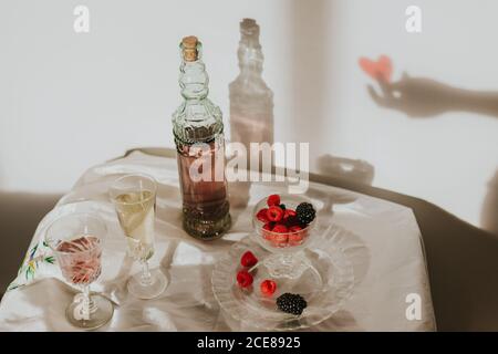 Von oben Flasche hausgemachten Rosenwein in Kristall gegossen Glas in der Nähe Champagner Trompete und serviert mit Teller frisch Himbeeren und Brombeeren gegen weiße Wand mit Handschatten Stockfoto