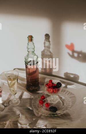 Von oben Flasche hausgemachten Rosenwein in Kristall gegossen Glas in der Nähe Champagner Trompete und serviert mit Teller frisch Himbeeren und Brombeeren gegen weiße Wand mit Handschatten Stockfoto