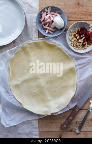 Draufsicht auf dünnen runden Blätterteig mit Gabelmarkierungen Auf Pergament neben Schalen mit Prosciutto Scheiben und Champignon mit Pinienkerne und sonnengetrocknete Tomaten für Quiche Stockfoto