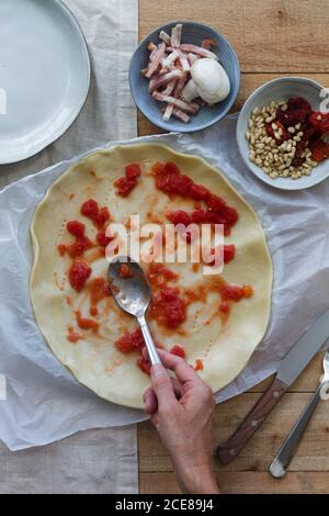 Draufsicht der Ernte anonyme kochen mit Löffel beim Putting Tomatensauce auf rohem Blätterteig in der Nähe von Schüsseln mit Speck Scheiben und Pinienkerne für Quiche während des Kochvorgangs Stockfoto