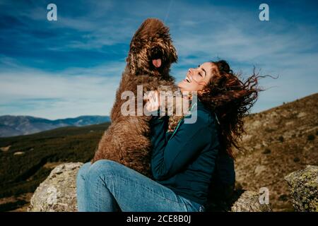 Seitenansicht der fröhlichen Besitzerin, die lacht und spielt Süße Labradoodle während der Rast in Puerto de la Morcuera Berge Der Himmel in Spanien ist bewölkt Stockfoto