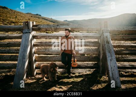 Ganzkörpermensch mit Ukulele und Labradoodle auf Holz gelehnt Barriere während der Rast an sonnigen Tag in Puerto de la Morcuera Berge in Spanien Stockfoto