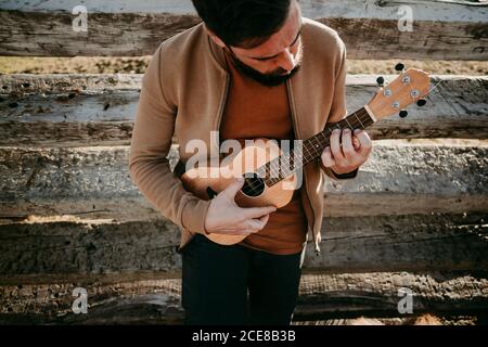 Von oben fokussierter Mann mit Ukulele, die auf Holzbarriere gelehnt ist Während Sie sich an sonnigen Tagen in Puerto de la Morcuera ausruhen Berge in Spanien Stockfoto