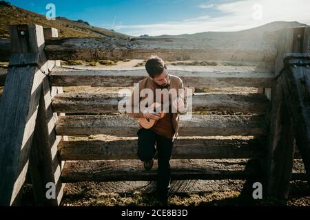 Von oben voller Körper des fokussierten Mannes mit Ukulele gelehnt Auf Holzbarriere, während Sie sich an sonnigen Tagen in Puerto ausruhen De la Morcuera Berge in Spanien Stockfoto