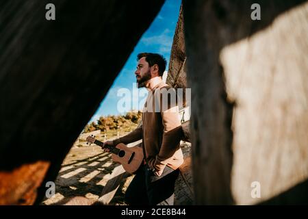 Von unten Seitenansicht des fokussierten Mannes mit Ukulele und Labradoodle lehnt sich an Holzbarriere, während er sich an sonnigen Tagen ausruhte In den Bergen von Puerto de la Morcuera in Spanien Stockfoto