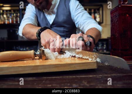 Cropped unkenntlich professionelle Barkeeper Schneiden Eiswürfel mit Messer auf Holzbrett während der Zubereitung eines Cocktails in der Bar Stockfoto