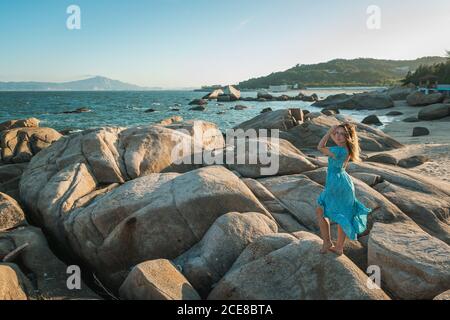 Ganzer Körper von jungen barfüßigen weiblichen Touristen in hellblau Kleid stehen auf riesigen Stein am Sandstrand und genießen Malerischer Blick auf das Meer bei Sonnenuntergang Stockfoto